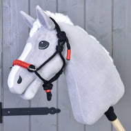 Hobby Horse LarDen Parelka Jiskra na šedém plyšovém Hobby Horse s bílou hřívou a černo-červenou parelkou, nasazeném na dřevěné tyči před šedou dřevěnou stěnou.