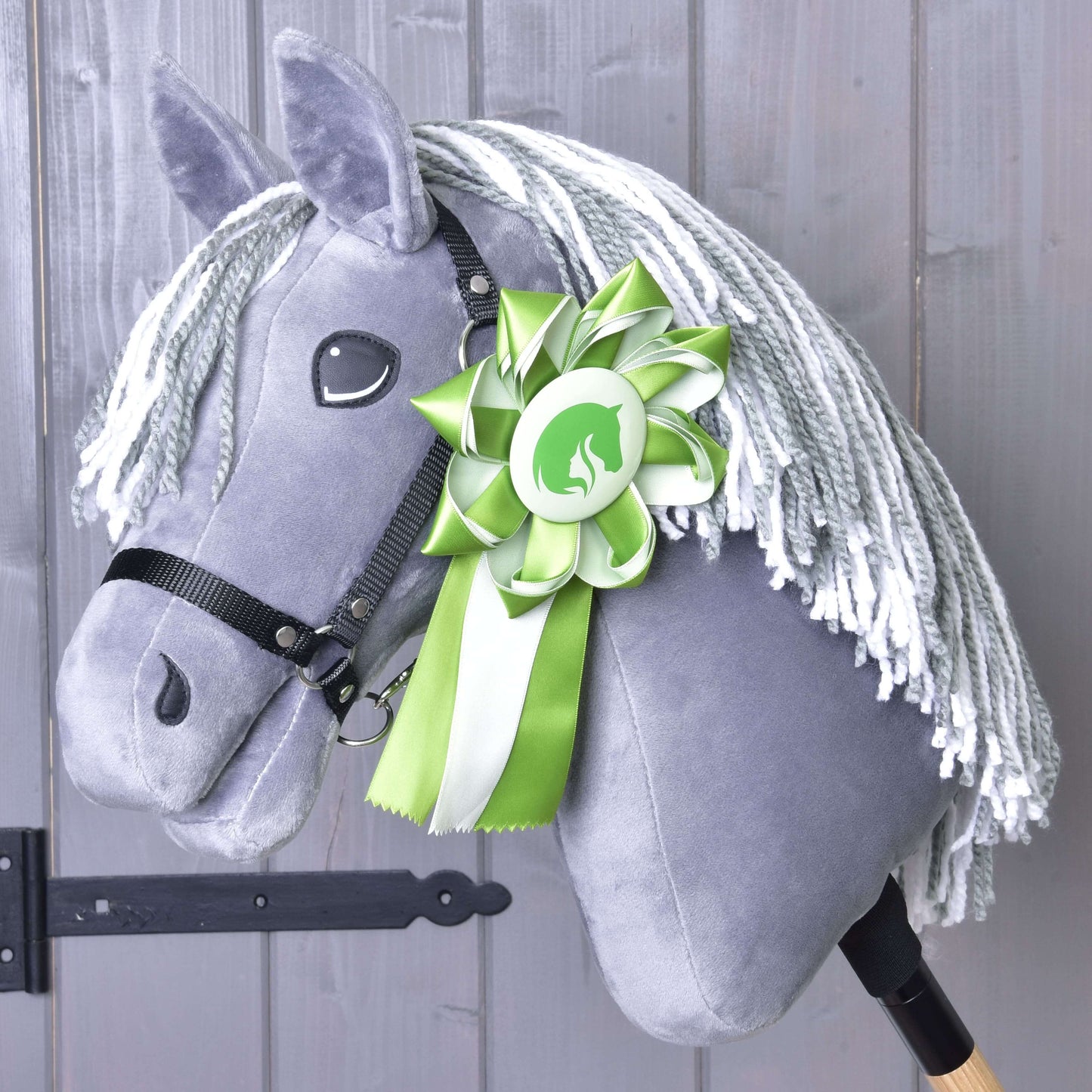 Hobby Horse Kokarda Zelená NEW od Hobby Horse LarDen s šedým plyšem, bílou přízovou hřívou, černou uzdečkou a zelenobílou kokardou připnutou na krku, zobrazený na dřevěném pozadí.