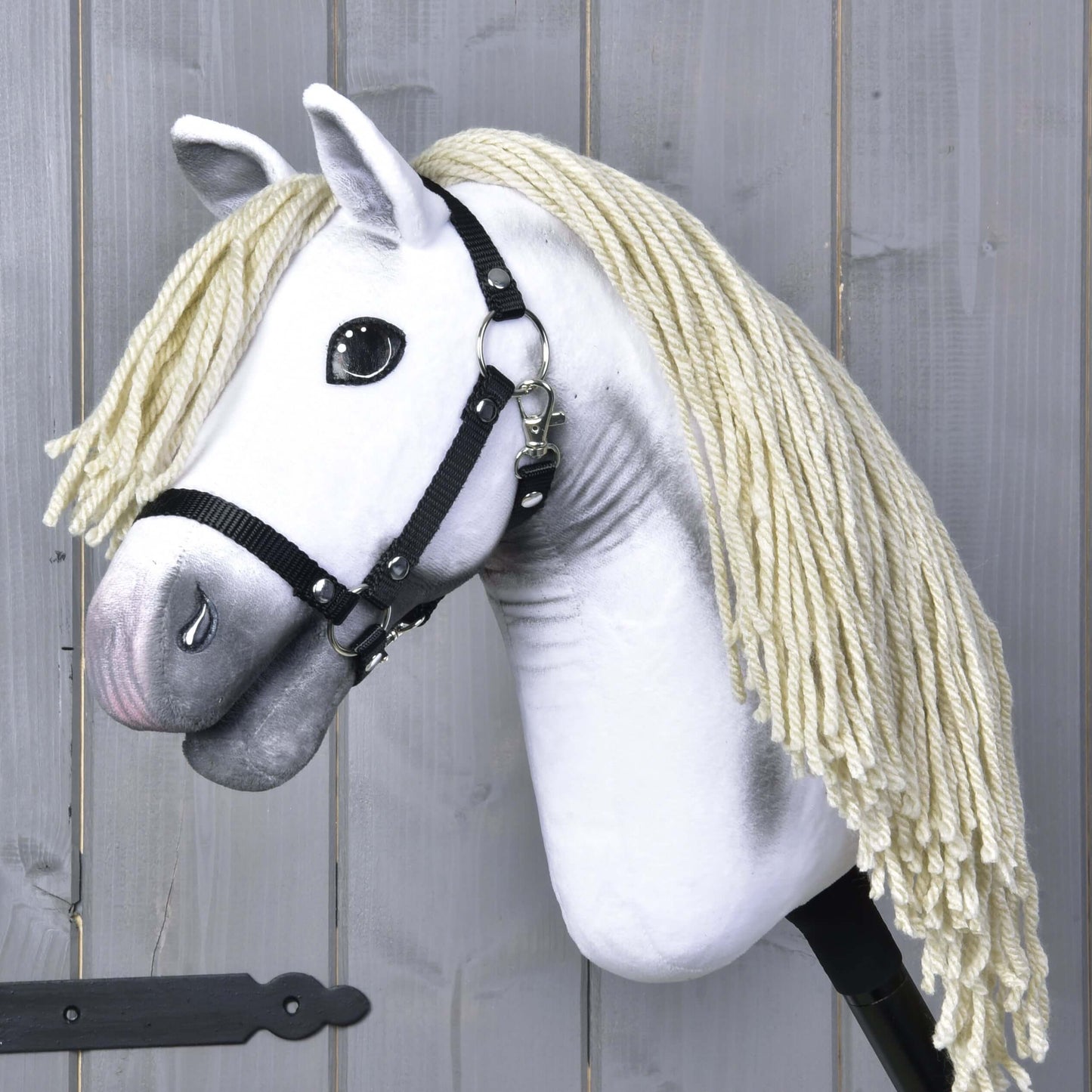 Hobby Horse Little Lady s černou ohlávkou od Hobby Horse LarDen má bílé tělo a dlouhou krémově zbarvenou hřívu z příze, stojí před dřevěnými panely a odráží eleganci drezury Pura Raza Española.