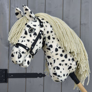 Hobby Horse Lord Valentine s černou ohlávkou od Hobby Horse LarDen, s plyšovou strakatou srstí, dlouhou krémovou hřívou a knoflíkovým okem, nasazený na dřevěné tyči před šedými dřevěnými panely.