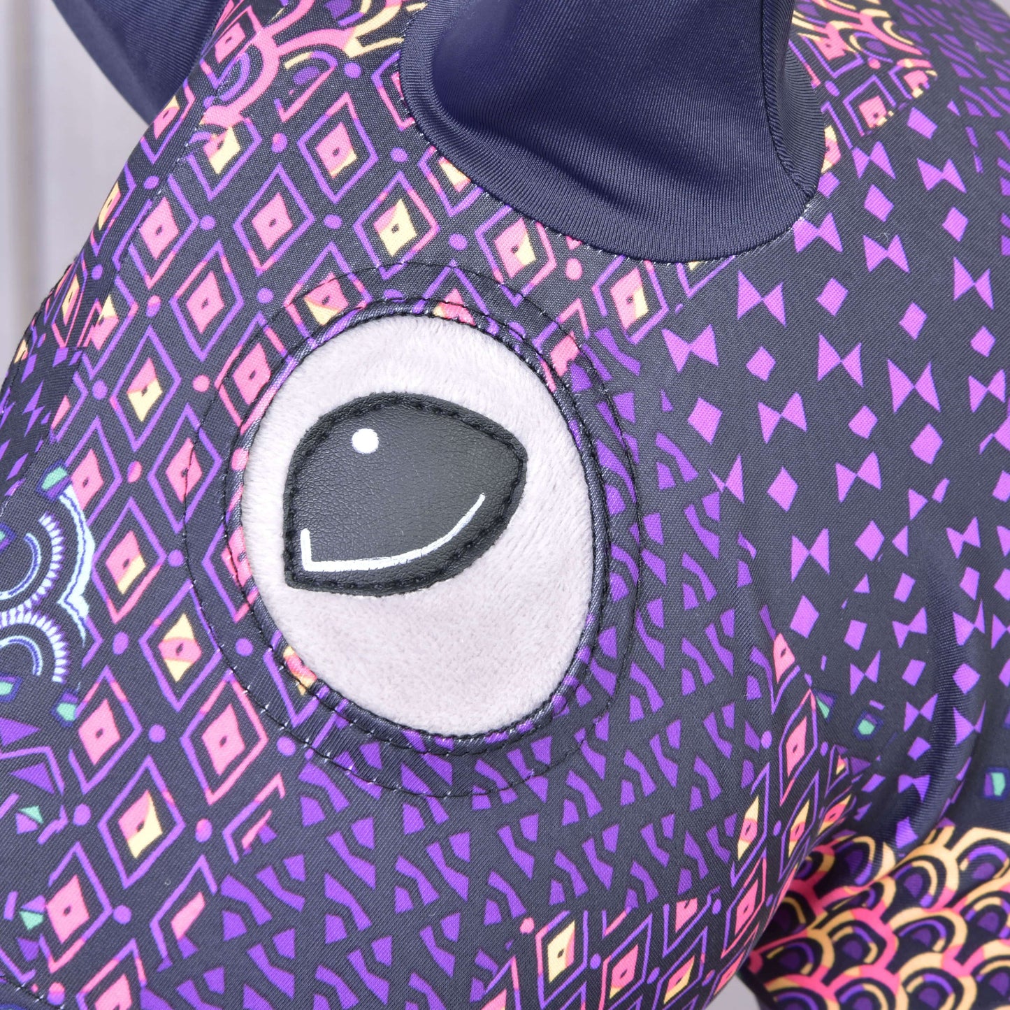 Detail ochranného obleku Hobby Horse ochranný oblek Mandala (Lycra) od Hobby Horse LarDen, zobrazující velké vyšívané oko a fialovou látku s barevnými geometrickými a kosočtvercovými vzory; nahoře je vidět tmavé ucho.