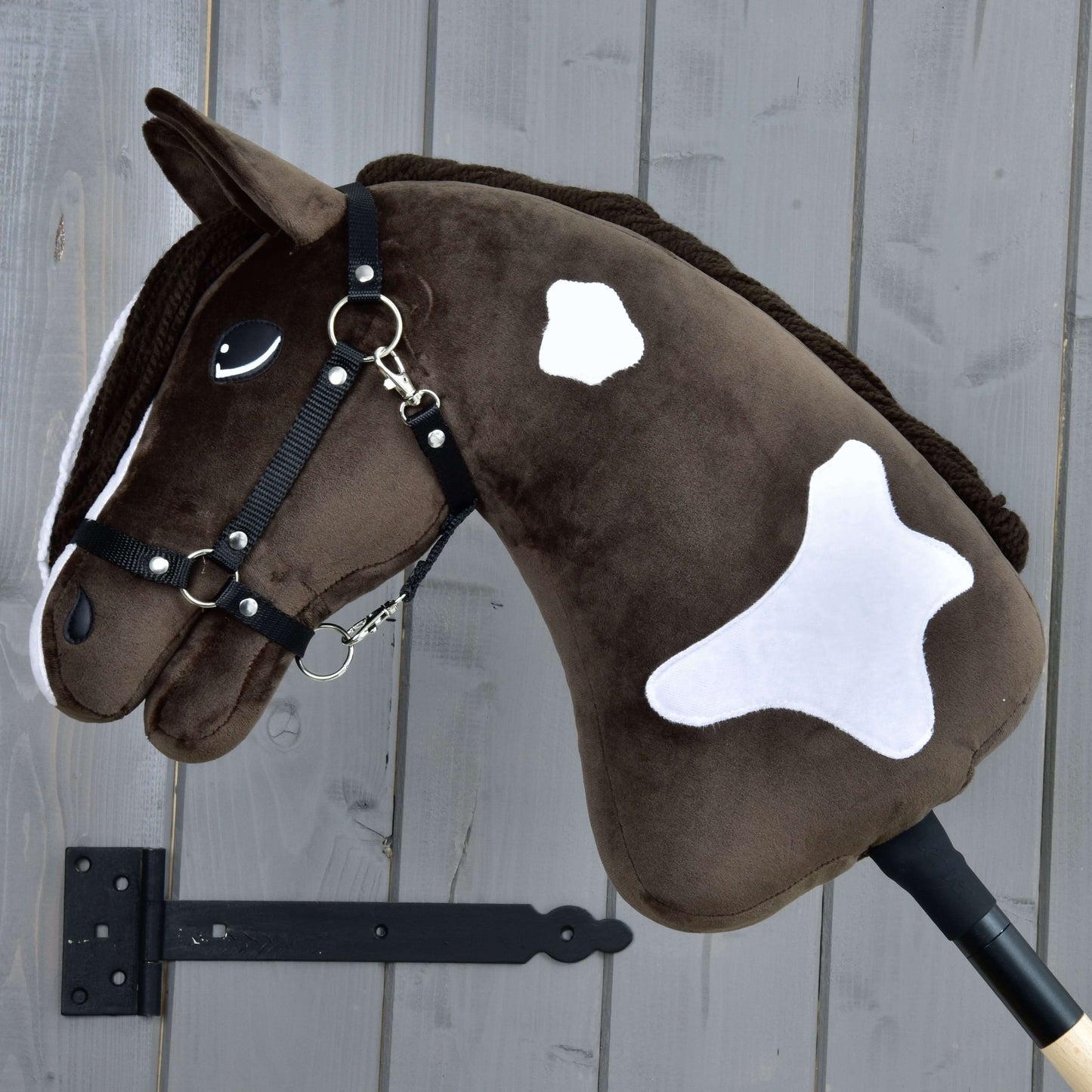 Hobby Horse Monte s černou ohlávkou od Hobby Horse LarDen je hnědobílý plyšový Hobby Horse inspirovaný American Paint Horse, s přátelským výrazem a bílými skvrnami, upevněný na šedé dřevěné stěně.