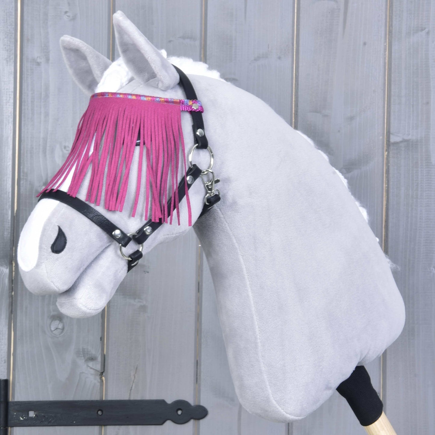 Plyšový Hobby Horse LarDen má na sobě Ohlávka černá s pink třásněmi a stojí před šedým dřevěným pozadím; má bílý kožich a stylový čelenkový řemínek s růžovými třásněmi.