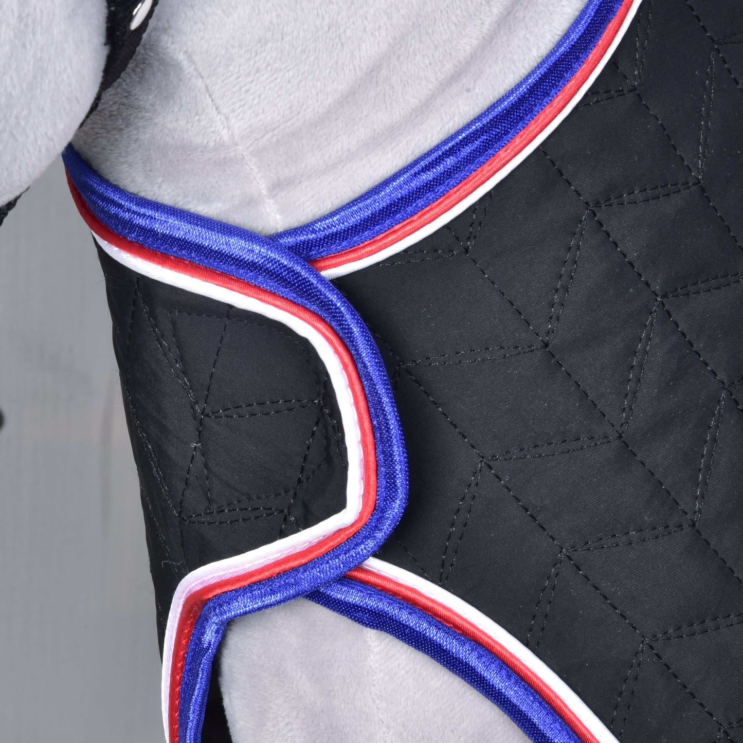 Detail černé prošívané látky s červeno-bílo-modrým lemováním, obepínající šedý povrch — pravděpodobně převozní deka Luxury od Hobby Horse LarDen, navržená pro vynikající pohodlí a ochranu během přepravy.