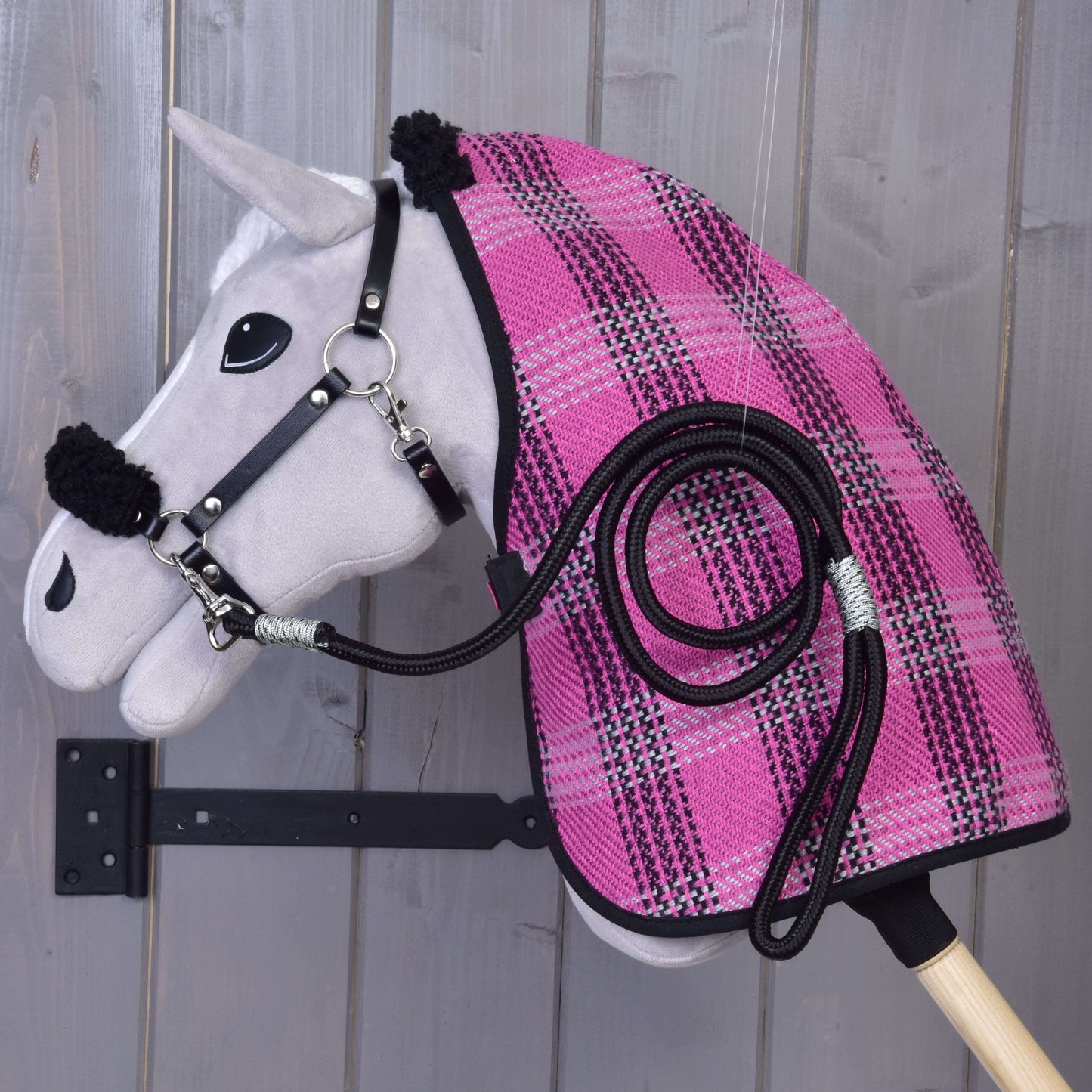 Šedý Hobby Horse ze setu „Set Harmony Pink“ od Hobby Horse LarDen je zavěšený na dřevěné stěně a má na sobě růžovo-černou kostkovanou stájovou deku, černou ohlávku a vedle sebe ladící stočené vodítko.