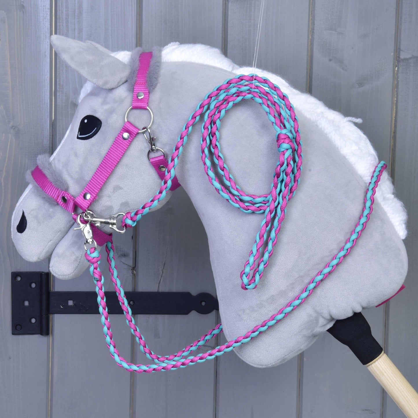 Set Perfect Combination Pink od Hobby Horse LarDen, šedý plyšový Hobby Horse s růžovou a tyrkysovou ohlávkou s kožešinovým polstrováním a otěžemi, visí na nástěnném háčku před šedým dřevěným pozadím.