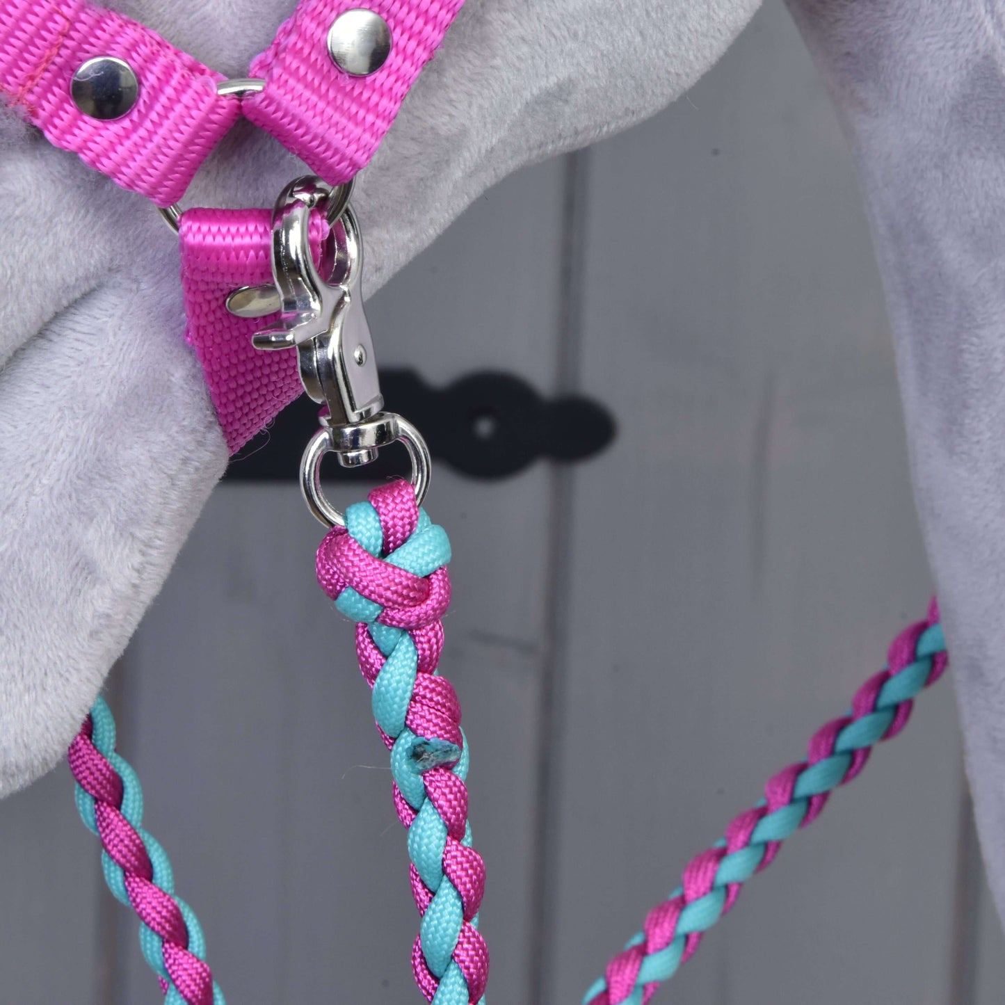 Detailní záběr na Set Perfect Combination Pink od Hobby Horse LarDen – růžový postroj a růžovo-tyrkysové pletené vodítko připnuté ke kroužku na šedém povrchu – ideální pro fanoušky stylových doplňků. V pozadí je vidět rozmazané světlé dřevo.