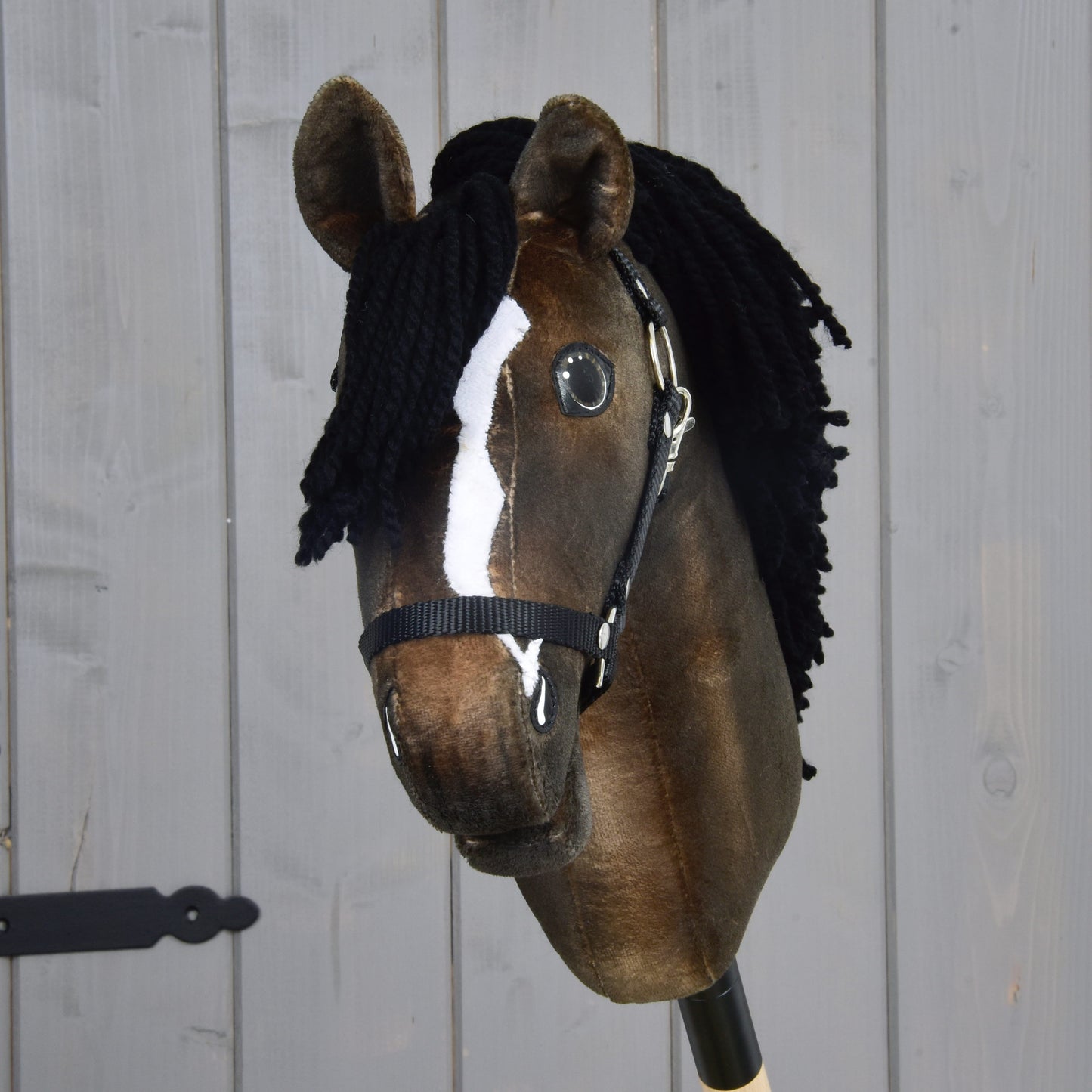 Hobby Horse Sundance s černou ohlávkou od Hobby Horse LarDen s hnědou srstí, černou přízovou hřívou, výraznou bílou lysinou a černou ohlávkou, stylizovaný podle anglického plnokrevného hřebce. Na pozadí světlé dřevěné stěny tato tvorba oslavuje jedno z nejrychlejších koňských plemen.