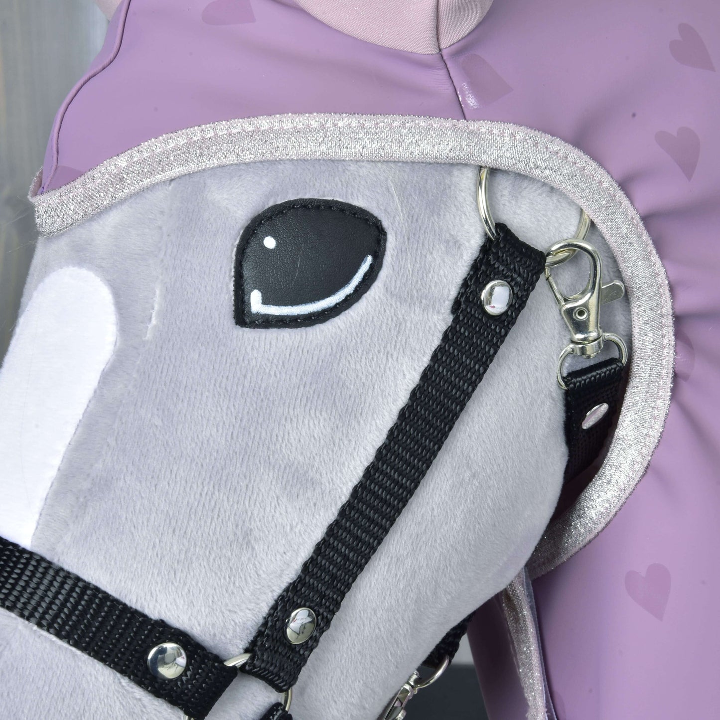 Detail plyšového šedého Hobby Horse Hobby Horse LarDen s černou uzdečkou, s doplňkem Pláštěnka Sweetheart, vedle osoby v levandulovém oblečení se srdíčkovým vzorem. Kůň má vyšívané usměvavé oko a třpytivý lem na krku.