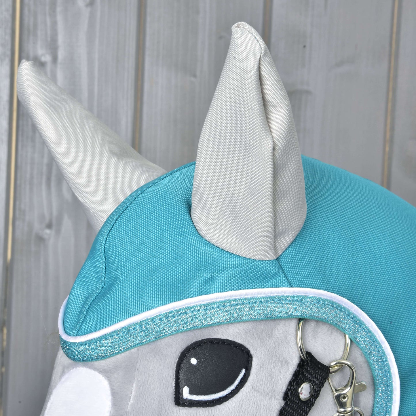 Detail hlavy plyšového koně Hobby Horse LarDen s Pláštěnkou Turquoise, se vztyčenýma ušima, třpytivým lemem a částí ohlávky – ideální pro hru za každého počasí na dřevěném pozadí.