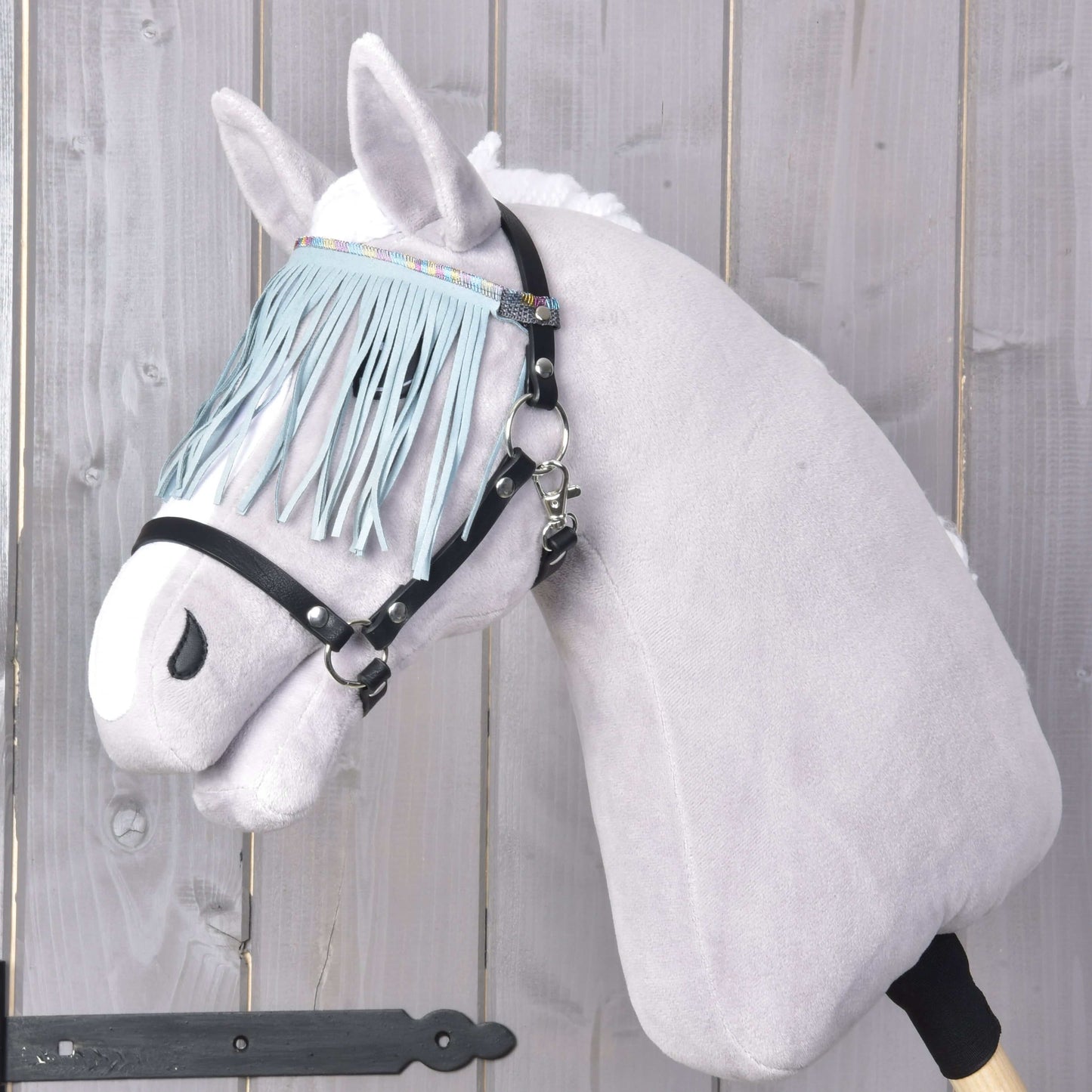Světle šedý model Hobby Horse LarDen s měkkým plyšovým obličejem a bílou hřívou je vybaven Ohlávka černá s třásněmi tyrkys light, včetně stylové černé uzdečky a tyrkysového čelenkového třásnění, na dřevěném pozadí.