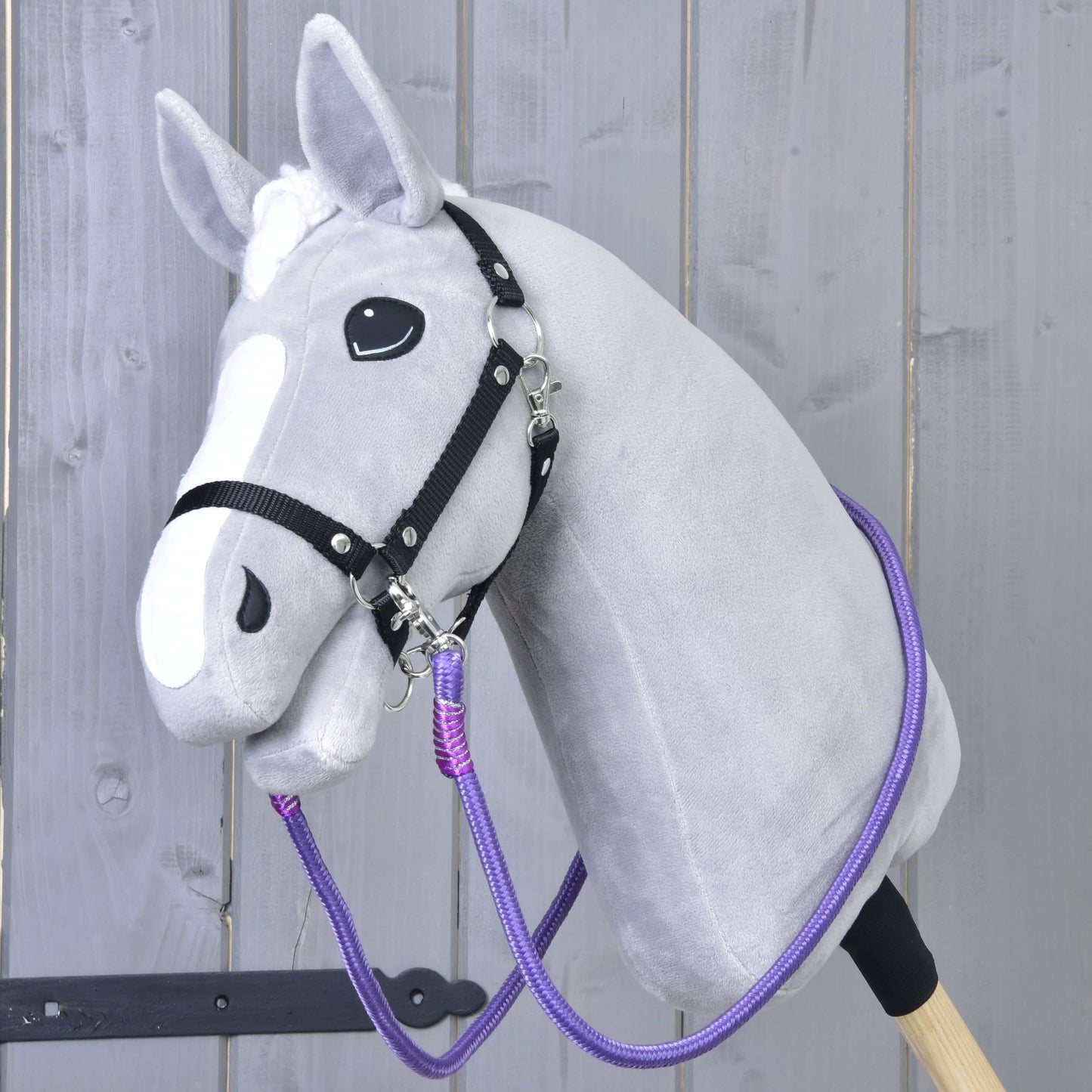 Hobby Horse LarDen „Otěže Viola“, šedý Hobby Horse s bílou lysinou, černou uzdečkou a výraznými ručně vyráběnými otěžemi, s plyšovou hlavou na pevné tyči, zobrazený před světlým dřevěným plotem.