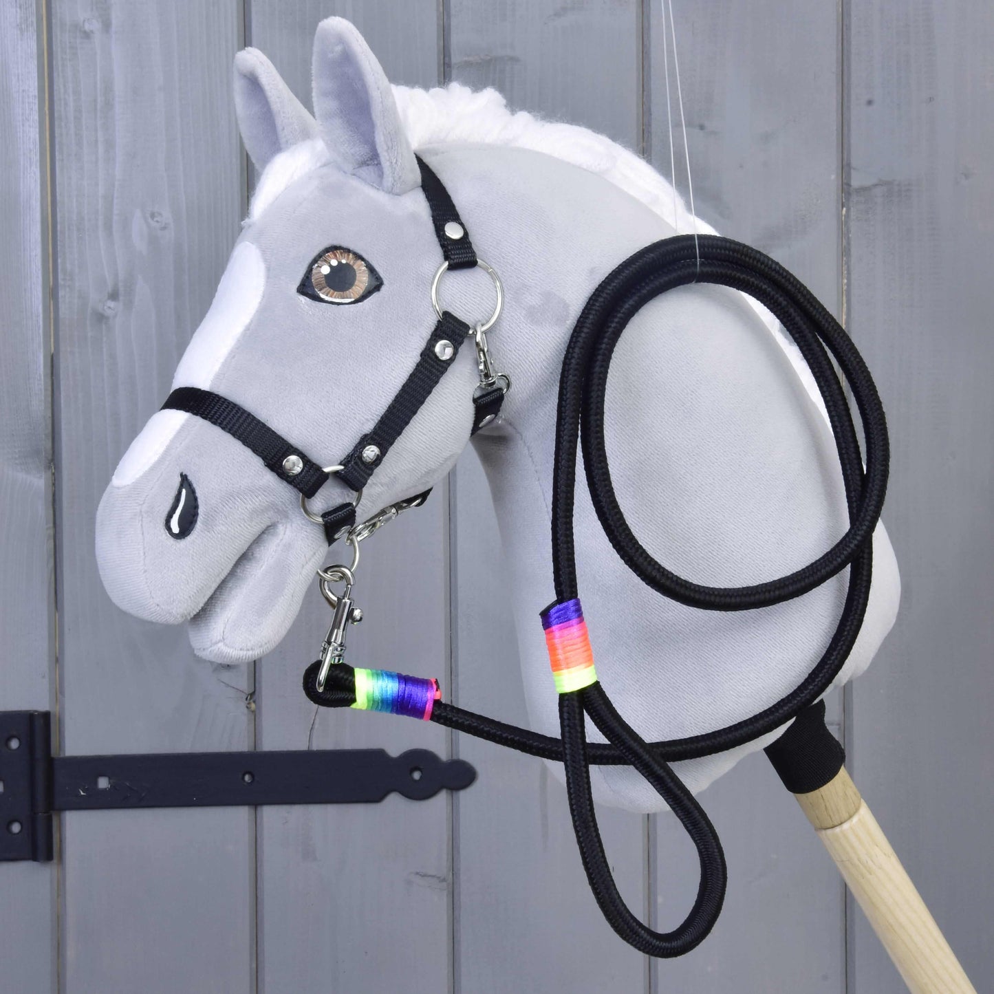 Hobby Horse - vodítko Rainbow od Hobby Horse LarDen na šedém Hobby Horse s bílou hřívou, černou uzdečkou, otěžemi a vodítkem s duhovou páskou kolem krku, vystaveném před dřevěným obložením.