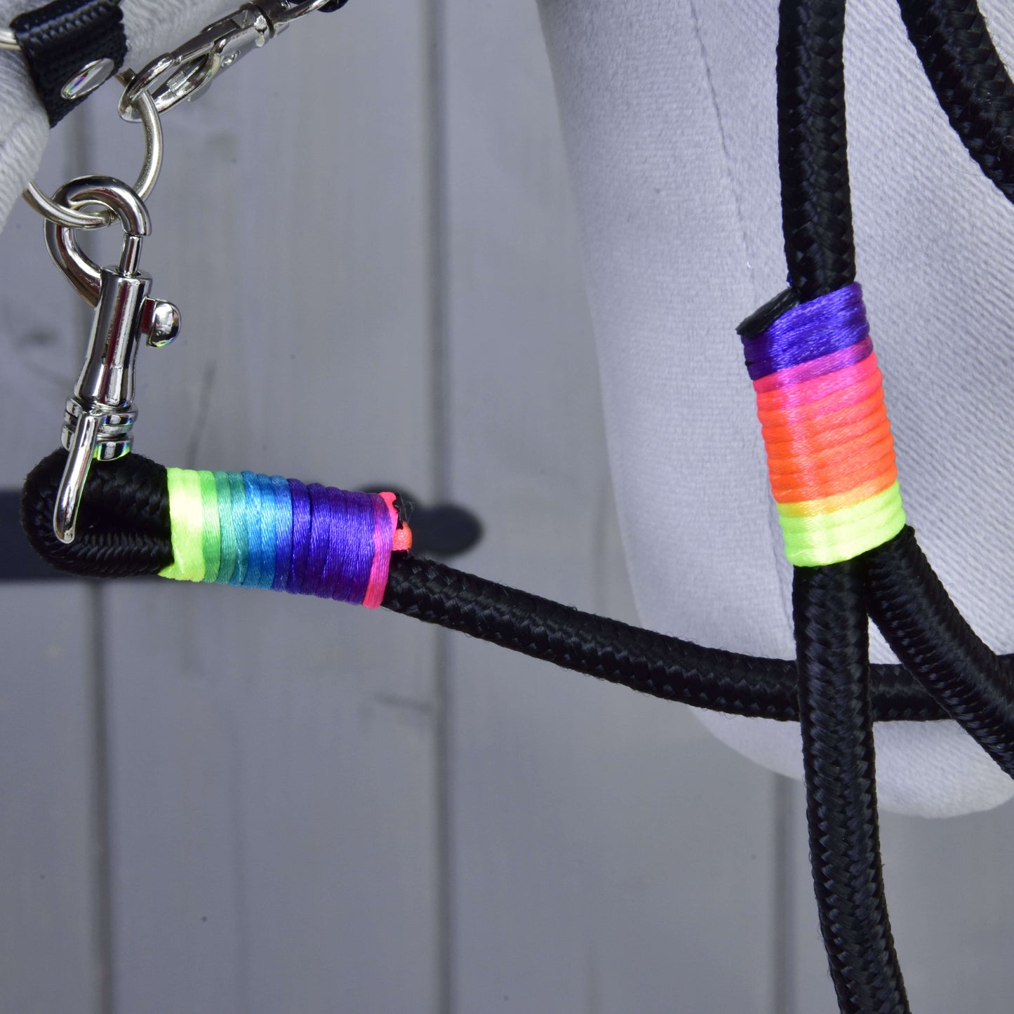 Detail Hobby Horse LarDen „Hobby Horse - vodítko Rainbow“, černého pleteného vodítka s omotávkami z neonově duhové nitě a stříbrnou karabinou, připevněného ke světle šedé látce, na dřevěném podkladu.