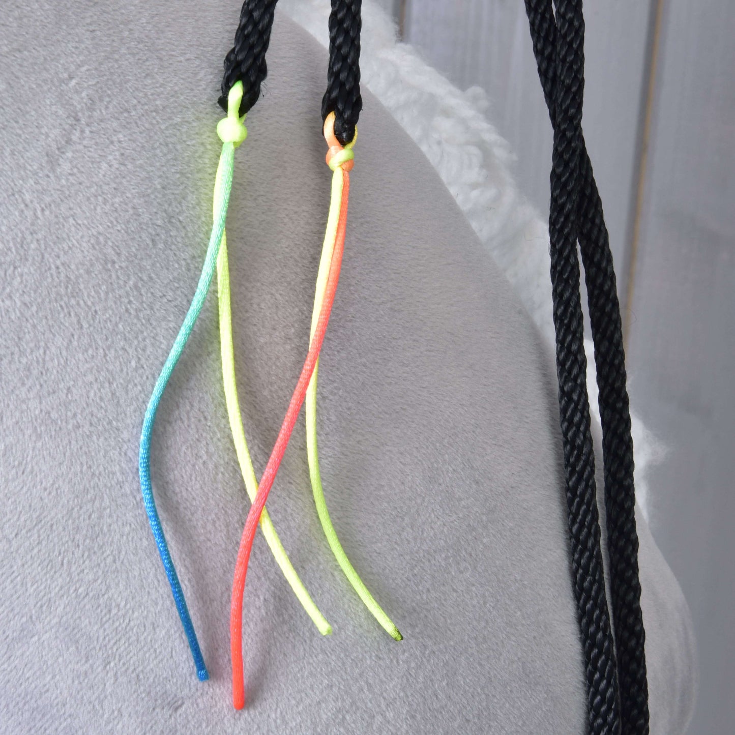 Detail Westernových otěží Texas rozdělených od Hobby Horse LarDen – černé pletené šňůry s neonově žlutými, modrými a oranžovými akcenty ze šňůrek v uzlech – položené na měkké světle šedé látce.