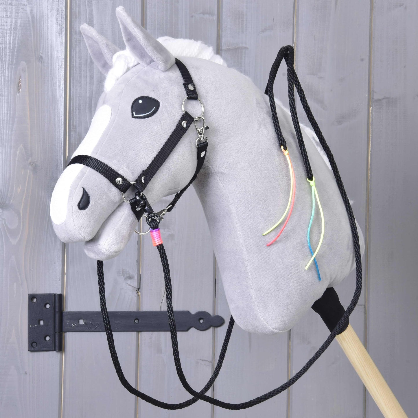 Hobby Horse LarDen "Westernové otěže Texas rozdělené", šedý plyšový koník s bílou hřívou, černou uzdečkou, dělenými westernovými otěžemi a barevnými stuhami na krku, nasazený na dřevěné tyči před šedými dřevěnými dveřmi.