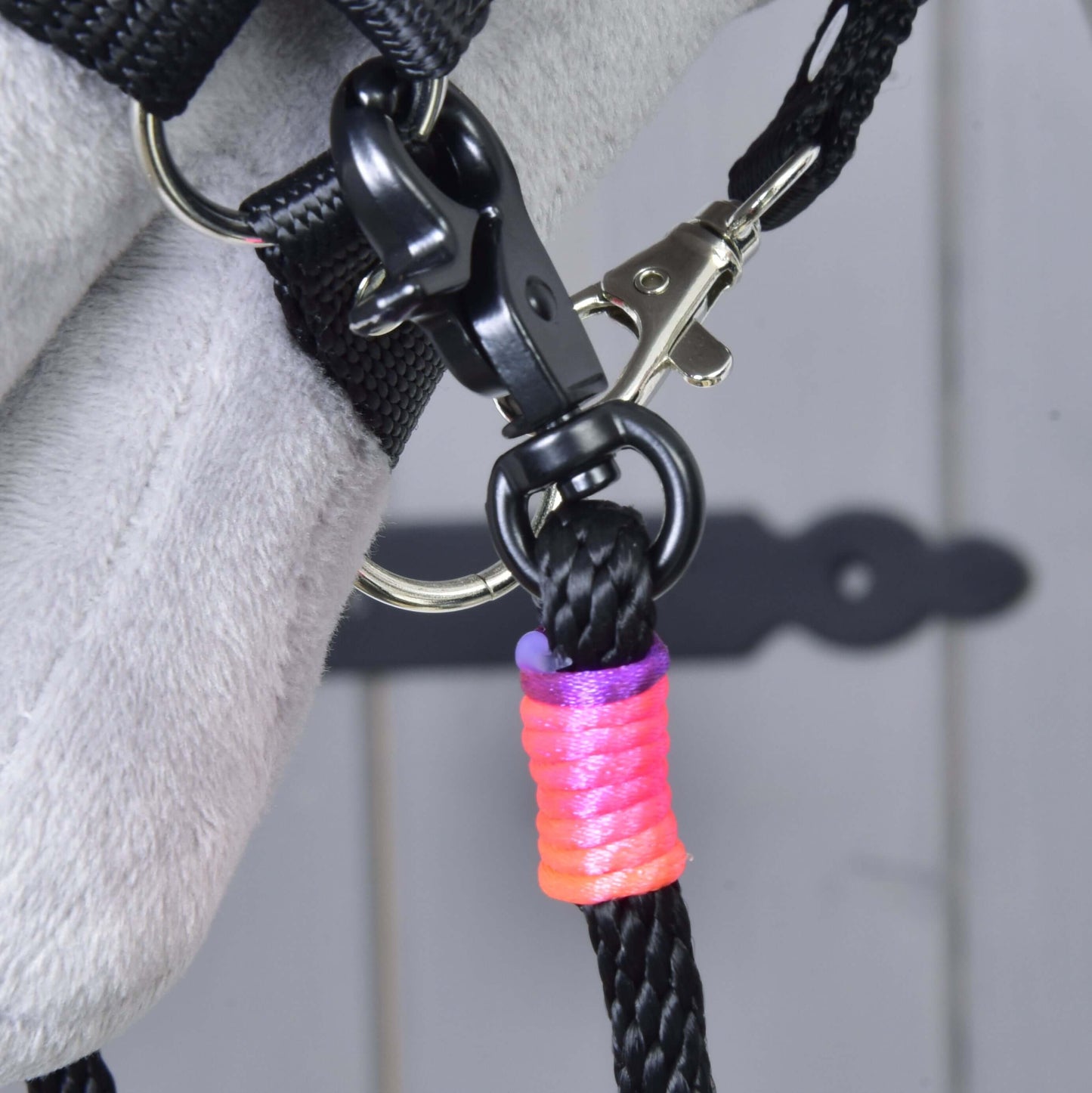 Detail Westernových otěží Texas rozdělených od Hobby Horse LarDen s černou kůží připnutou karabinami ke kovovému kroužku na šedém povrchu a s omotáním u karabiny růžovou, oranžovou a fialovou nití.