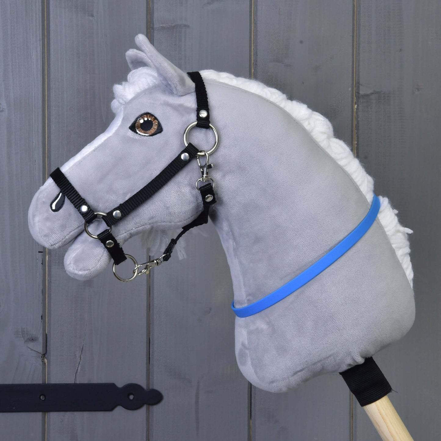 Hobby Horse nákrčník modrý (vel. S) od Hobby Horse LarDen na šedém plyšovém Hobby Horse s bílou hřívou, černou uzdečkou a modrofialovou stuhou na dřevěné tyči, před šedou dřevěnou stěnou.